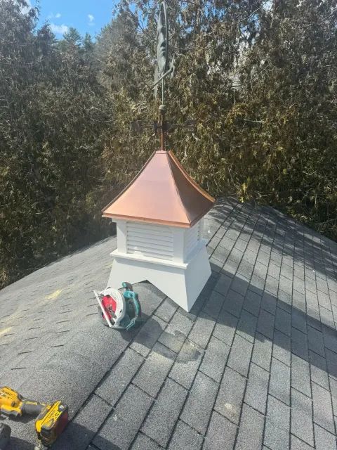 Copper-topped white rooftop cupola on a gray shingled roof with trees in the background.