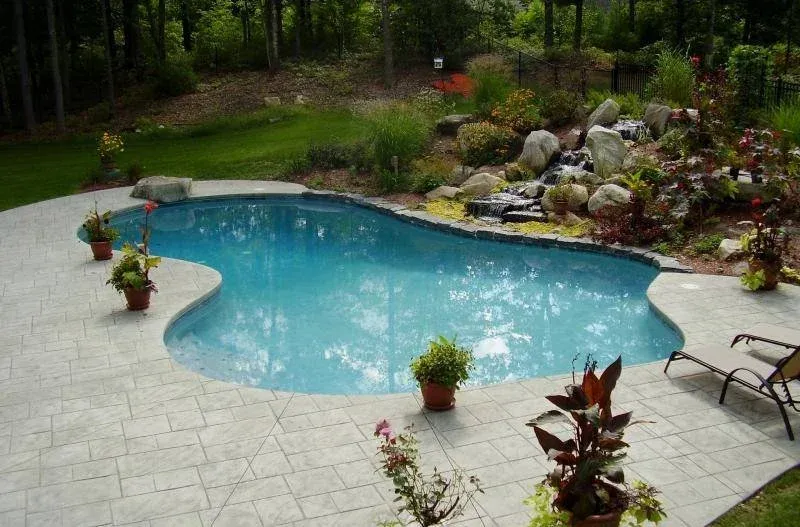 Backyard with a kidney-shaped swimming pool, surrounded by potted plants and lounge chairs, with a landscaped rock garden and trees in the background.