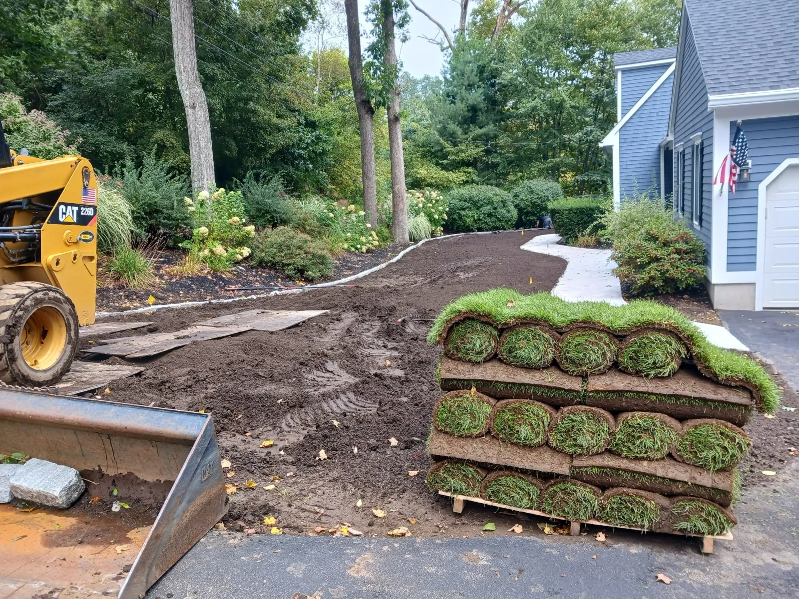 A pathway under construction in a residential yard, with a stack of sod rolls and gardening equipment visible. The pathway curves along a blue house with white trim, surrounded by trees and shrubs.