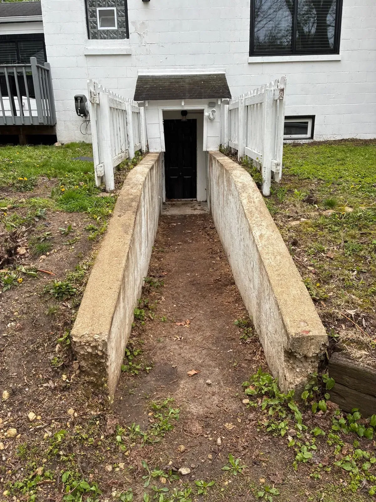 Narrow concrete ramp leading to a basement door on a white house, bordered by low retaining walls.