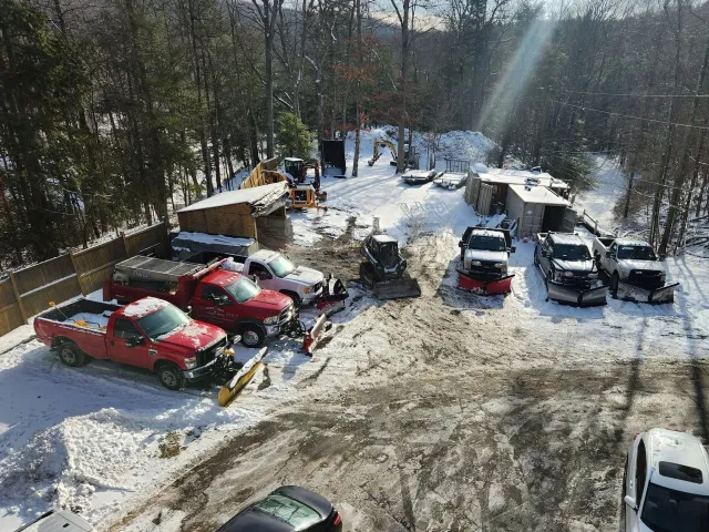 Snow-covered parking lot with several parked cars and trees in the background.