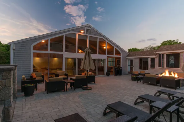 Outdoor patio with seating, fire pit, and umbrella, in front of a house with large windows at sunset.