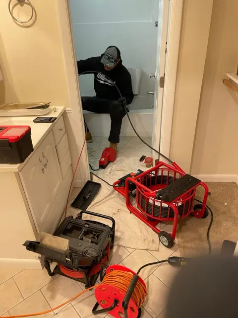 Person in a bathroom with cleaning equipment and tools scattered across the floor; robot-like devices are visible.