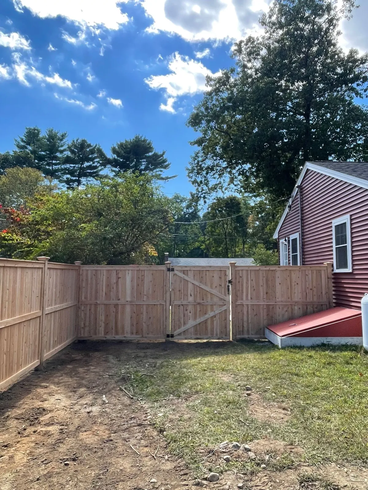 Wooden fence enclosing a backyard with a red house. A gate is in the center. Blue sky with white clouds.