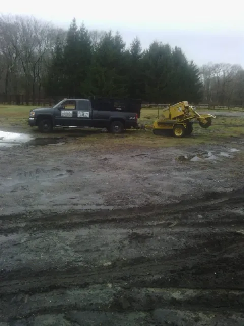 A black pickup truck with signs on the door is attached to a yellow portable sawmill machine, parked on muddy ground near water puddles, with a background of trees and an overcast sky.