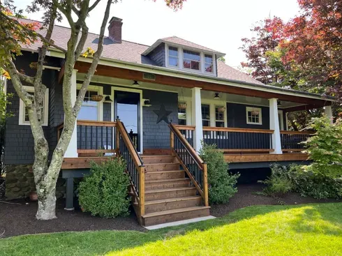 A house with a wraparound porch and steps, painted dark gray, with a star on the wall, and a green lawn.
