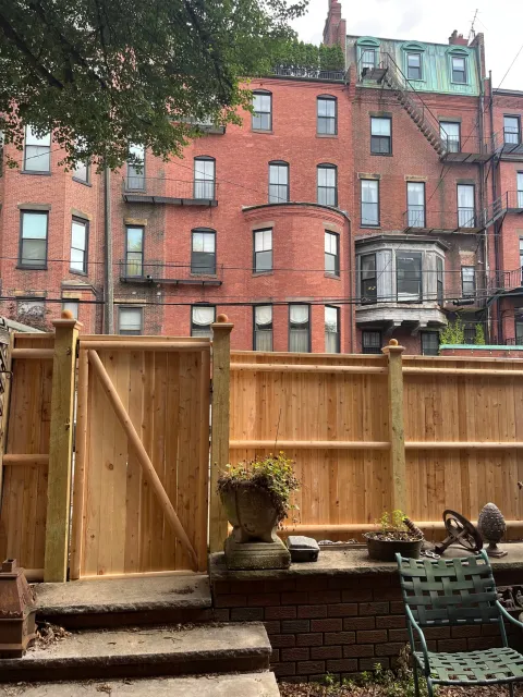Backyard scene with a wooden fence, potted plants, garden tools, and outdoor furniture, with an apartment building in the background.