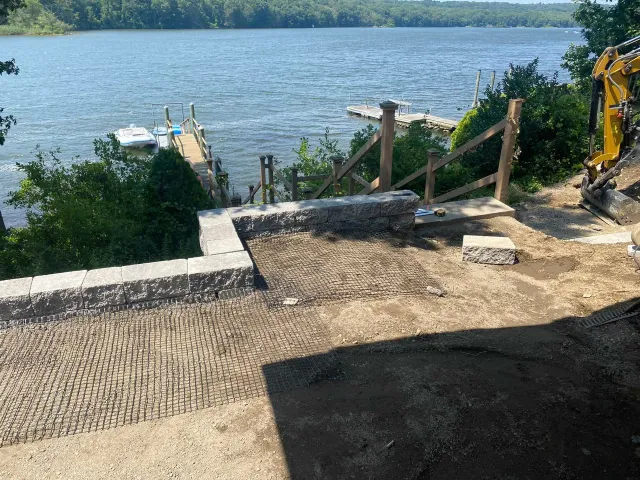 A stone retaining wall overlooks a lake with docks and a yellow excavator parked on the right side.