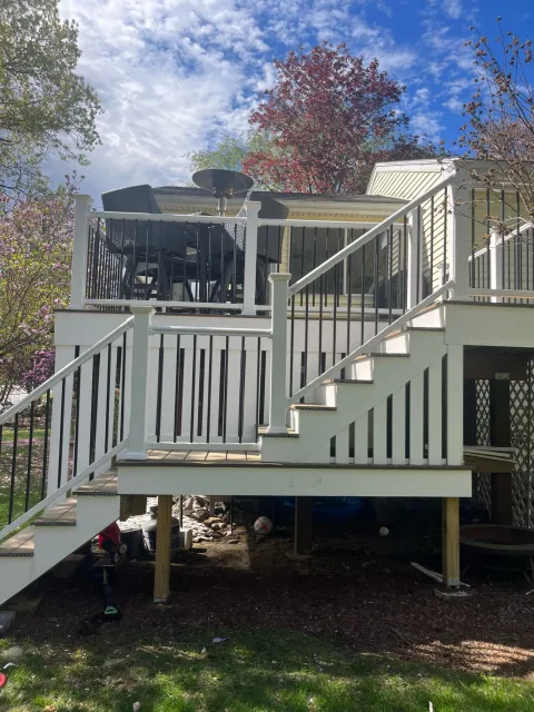 Outdoor deck with stairs, patio furniture, and umbrella heater, surrounded by trees under blue sky with clouds.