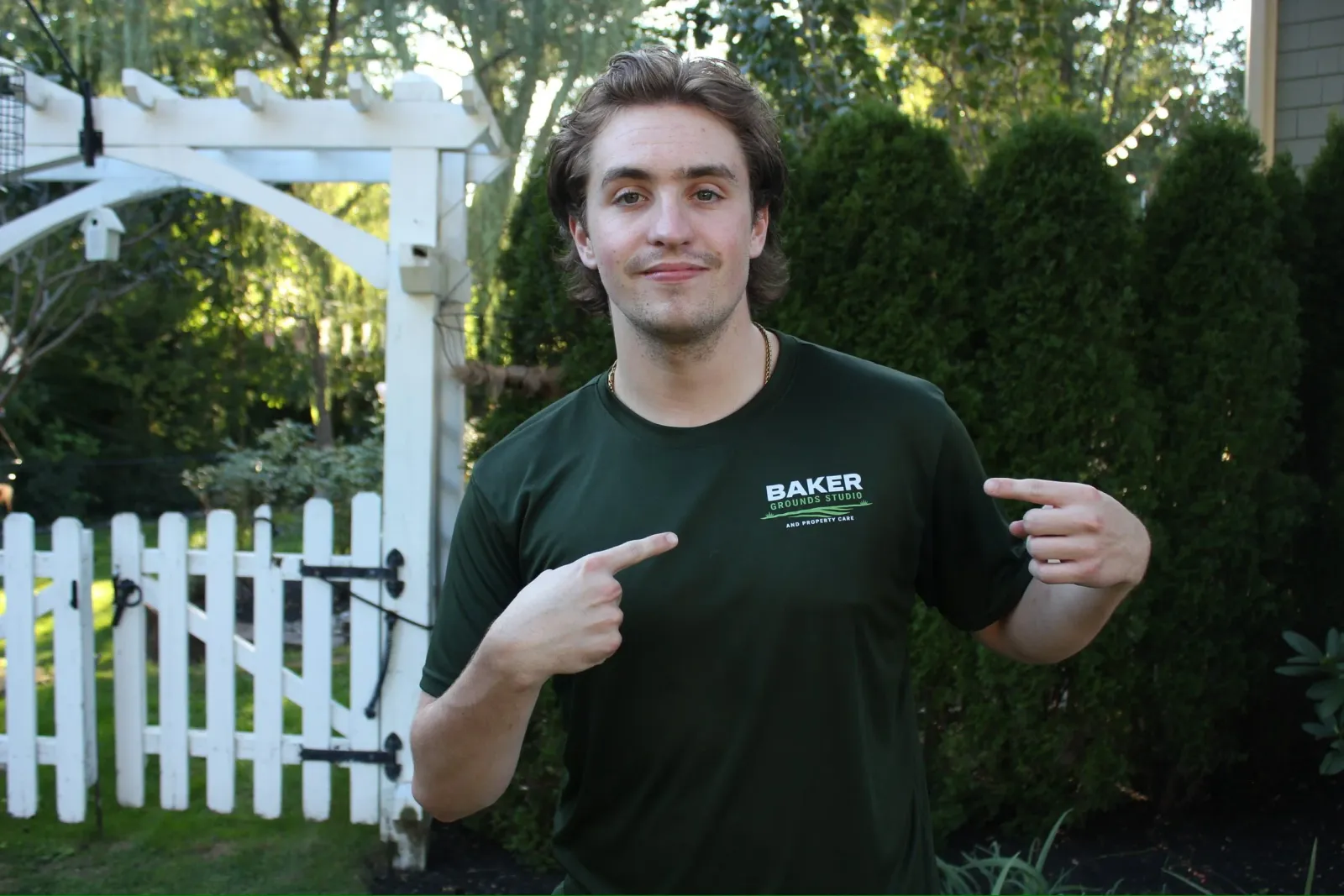 A young man with brown hair and a slight smile pointing to the logo on his black t-shirt, which reads 'Baker Grounds Studio and Property Care,' outdoors in a garden with trees, bushes, a white picket fence, and a white arched garden trellis in the background.