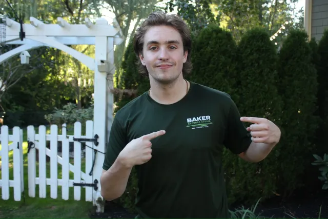 A young man with brown hair and a slight smile pointing to the logo on his black t-shirt, which reads 'Baker Grounds Studio and Property Care,' outdoors in a garden with trees, bushes, a white picket fence, and a white arched garden trellis in the background.