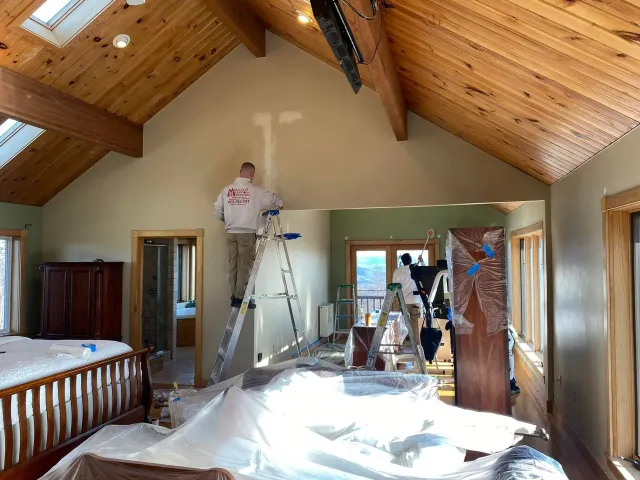 Workers on ladders painting a vaulted bedroom with a wood ceiling and plastic-covered floor.