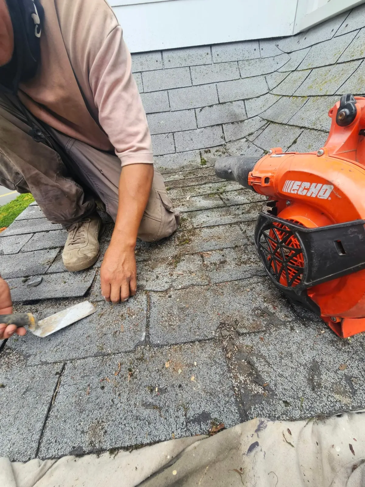 Worker installing gray roof shingles beside an orange saw on a rooftop