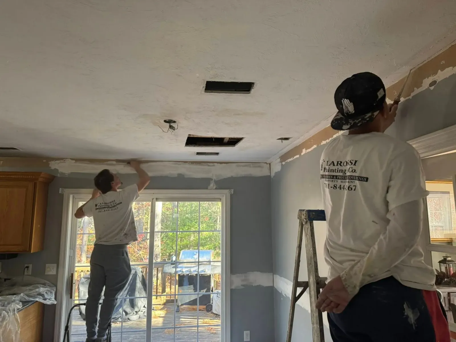 Two workers patching and painting a room ceiling near a window and ladder