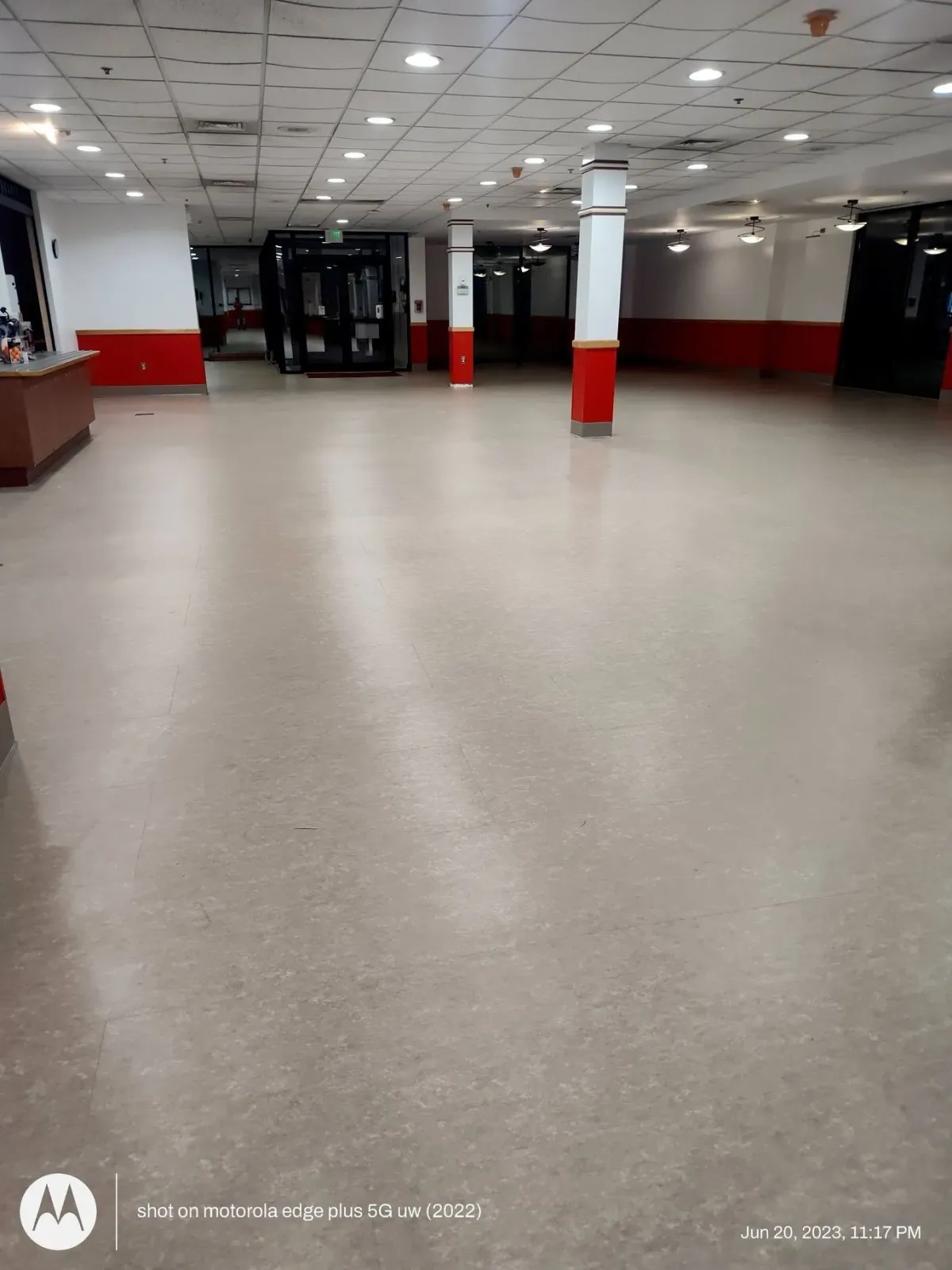 An empty commercial space with light-colored flooring, white support columns, and red wall trim under a drop ceiling.