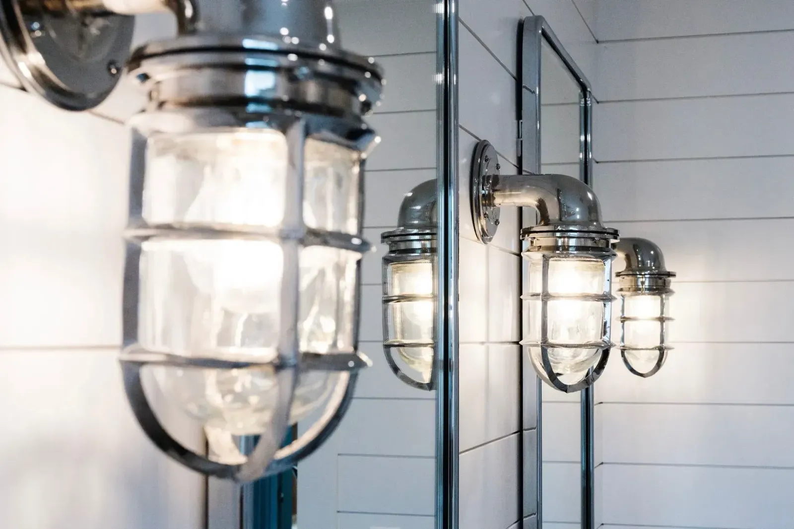 Three chrome industrial wall lights with glass cages on a white tiled wall