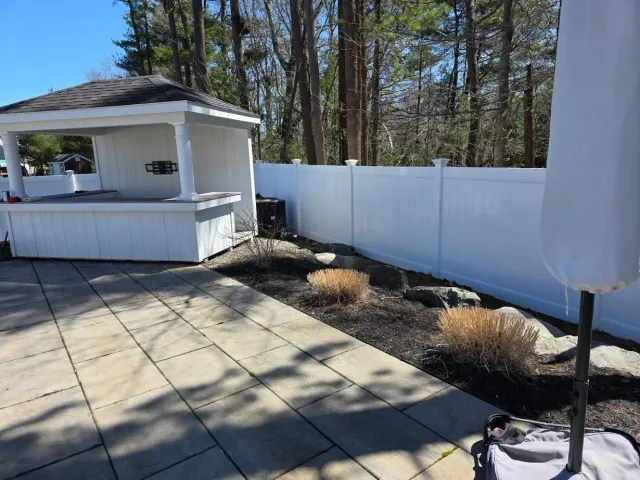 Patio with white gazebo, paver stones, and white fence beside a wooded yard