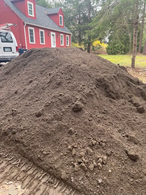 A large pile of dirt in a backyard with a red house in the background, trees, and a white van nearby.