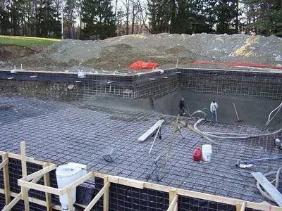 Construction site with a concrete foundation, rebar reinforcement, and workers preparing the area amid trees in the background.