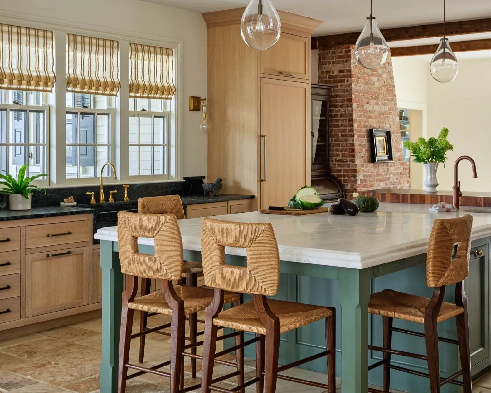A modern kitchen with a large central island with a white marble countertop and four rattan barstools. The kitchen has light wood cabinets, black countertops, and a brick fireplace. There are three large windows with striped shades, hanging glass pen