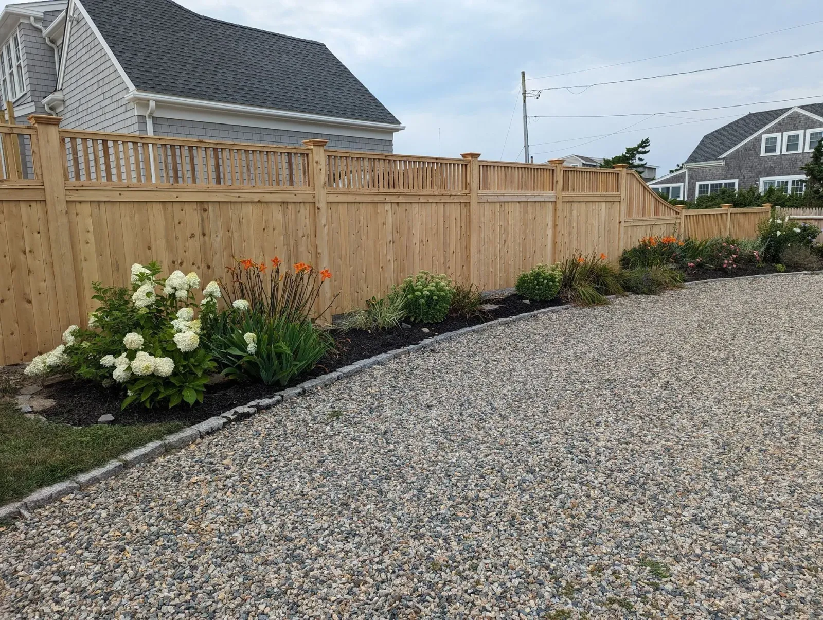 A light wood fence separates a gravel driveway from a garden with white hydrangea bushes and orange flowers.