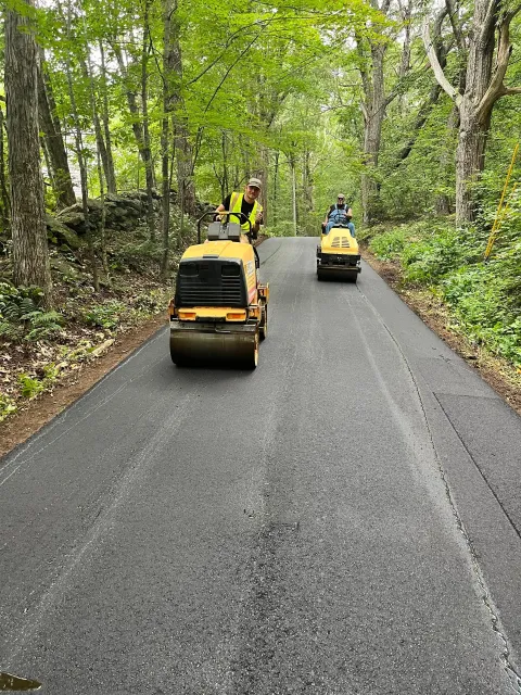 Two asphalt rollers compact fresh pavement on a tree-lined road.