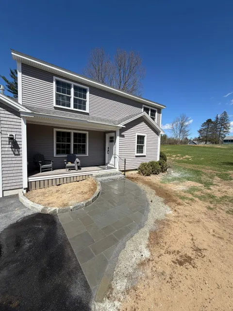 Gray house exterior with small porch, stone walkway, and bare yard under a blue sky.