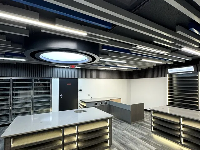Renovated gas station convenience store sales floor in Northford Connecticut with a black acoustic baffle ceiling with navy accents and linear LED strips, a circular illuminated ceiling feature, a glass-door reach-in cooler line, black slat wall panels, LED-lit island gondola displays with light quartz tops, a coffee and beverage service counter, and grey wide-plank commercial vinyl plank flooring