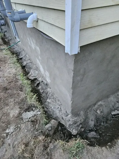 A gray concrete building foundation meets beige siding, showing exposed footing in a muddy trench alongside the house.