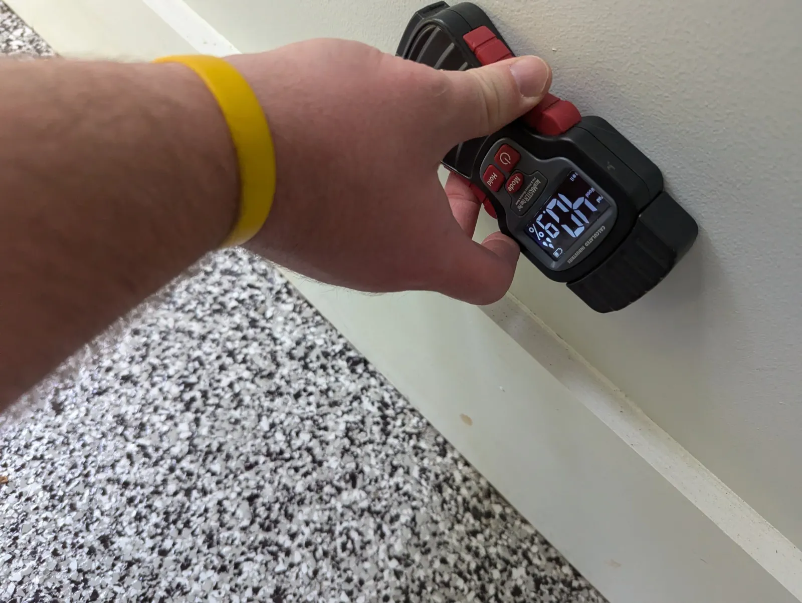 A person holding a digital remote control or device with a digital display showing the time 12:09, against a background of a textured speckled flooring and a plain white wall.