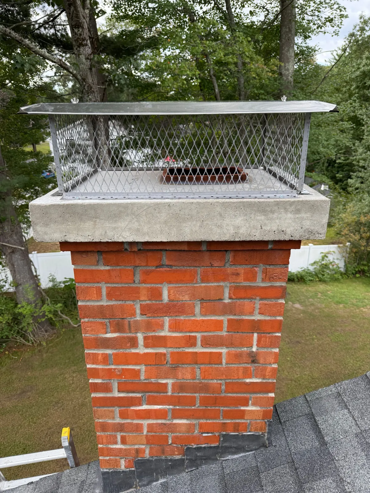 A brick chimney with a metal cage cap on top, located on a rooftop with shingles, surrounded by trees in the background.