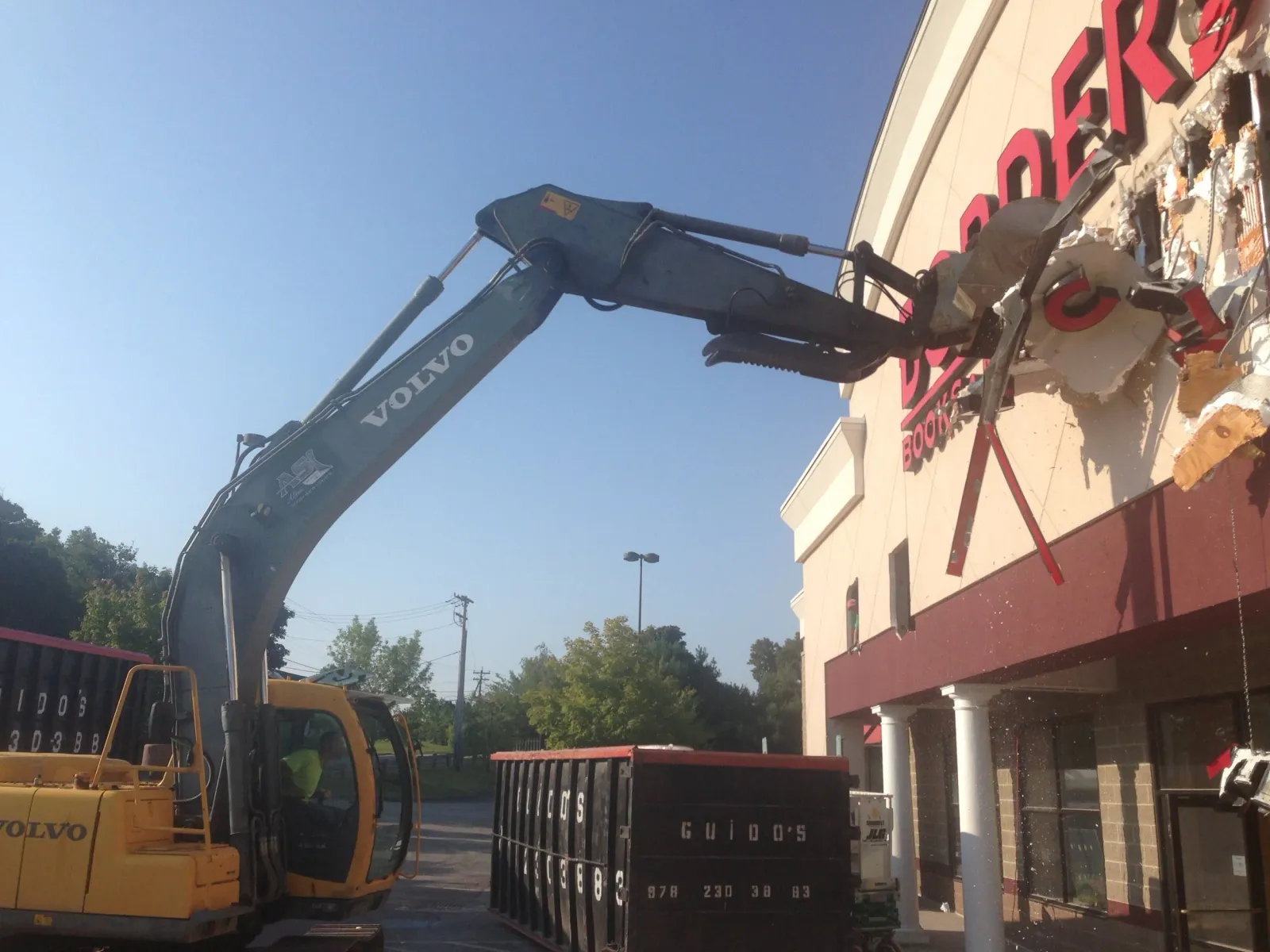 A construction scene with a Volvo excavator and a dump truck parked near a building with large pink letters on the wall. The building has a beige upper section and red lower section with white columns, and the scene is outdoors on a sunny day.