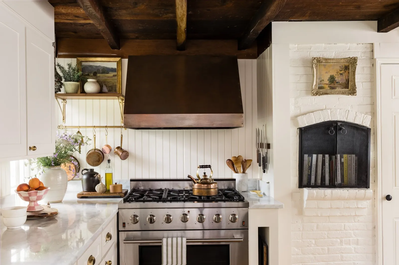 Rustic cottage farmhouse kitchen with stainless steel stove, wooden countertops, copper kettle and hood, white cabinets, potted plants, hanging pots, cutting boards, bowls with fruit, and a decorative wall niche with books and artwork.