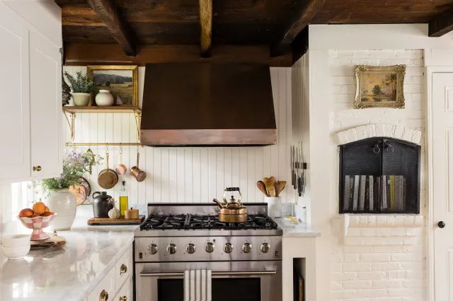 Rustic cottage farmhouse kitchen with stainless steel stove, wooden countertops, copper kettle and hood, white cabinets, potted plants, hanging pots, cutting boards, bowls with fruit, and a decorative wall niche with books and artwork.