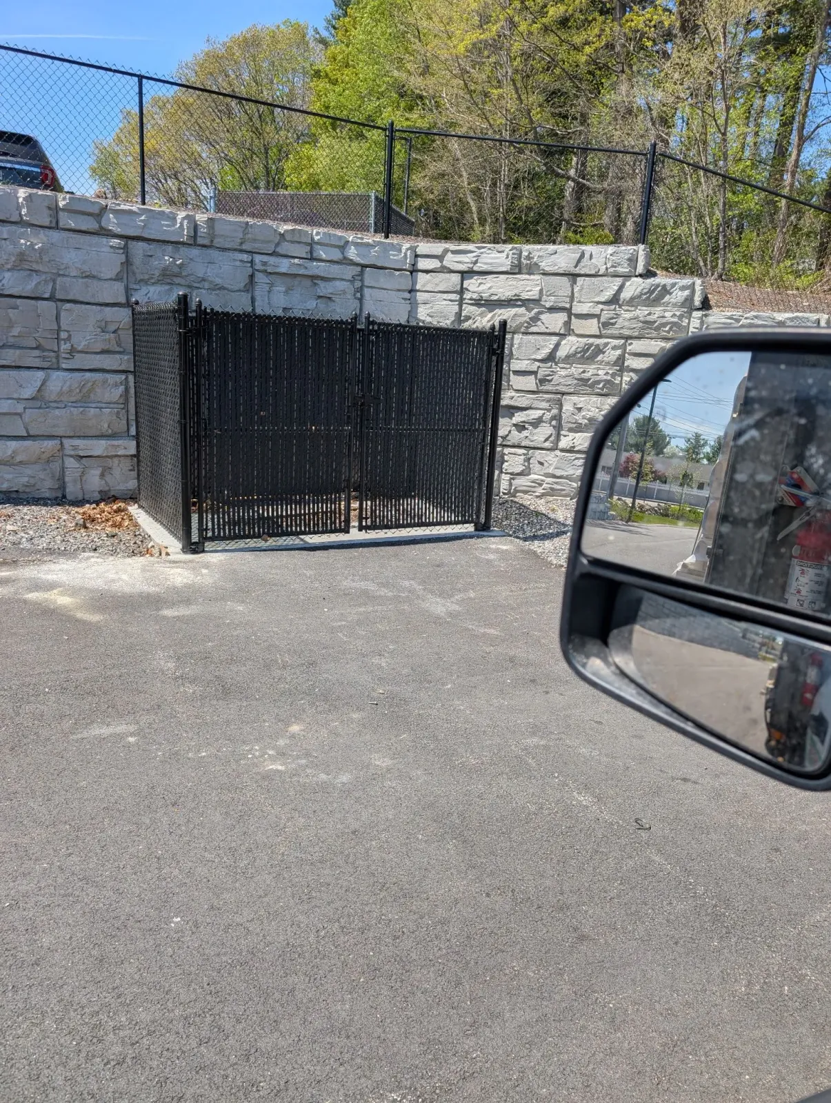 A black metal-fenced trash enclosure set into a stone retaining wall in an outdoor parking area.