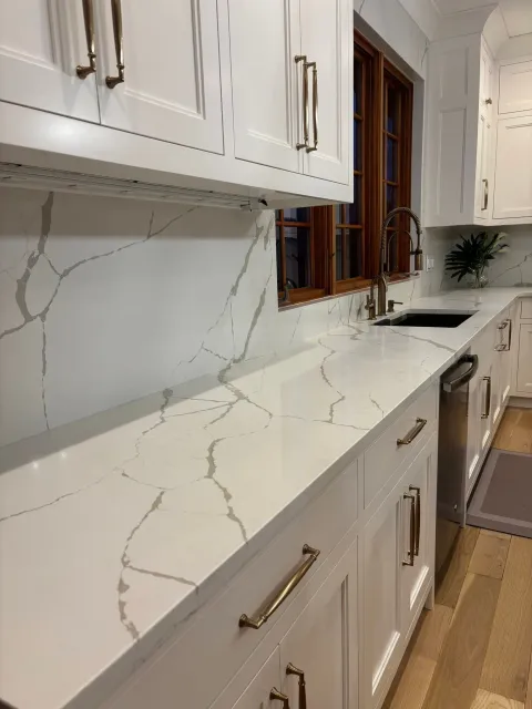 Modern kitchen counter with white marble-look countertop, white cabinets, brass handles, and a window above the sink.
