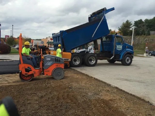 Workers operate a vibratory roller and a paver alongside a blue dump truck depositing asphalt at a construction site.