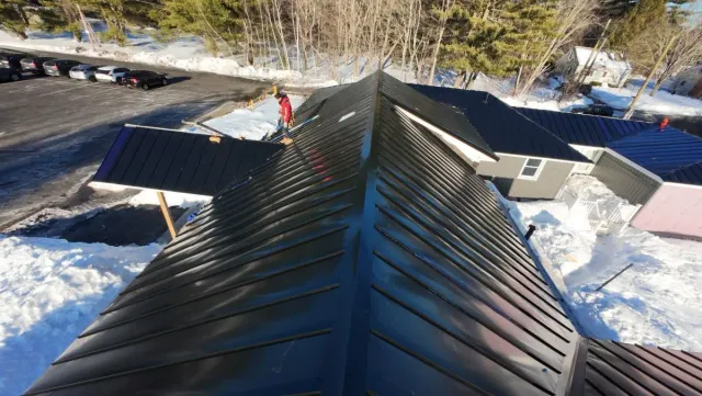 Snowy rooftop ridge with dark metal shingles and neighboring houses below on a sunny day