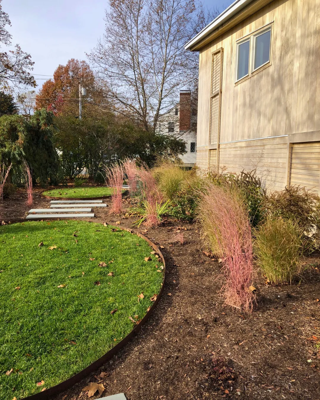 A modern garden with a curved, mulched flower bed featuring pink ornamental grasses. The bed borders a green lawn with several stepping stones. Trees with autumn foliage are in the background, next to a beige wooden building.