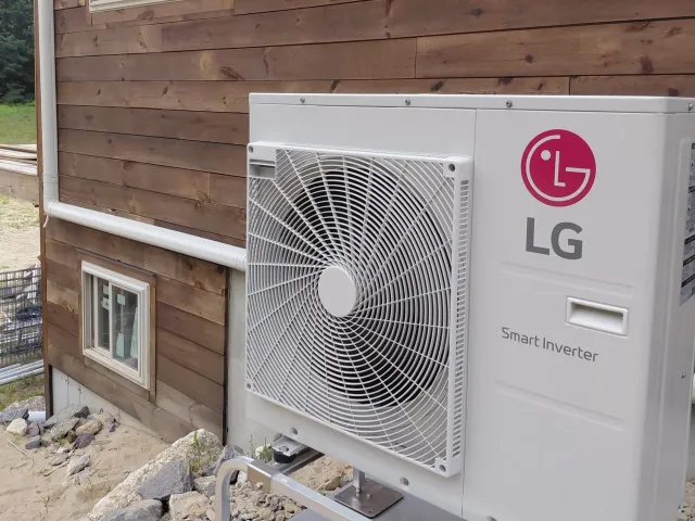 An outdoor LG air conditioning unit mounted on the exterior wall of a wooden house with visible piping and a small window below.