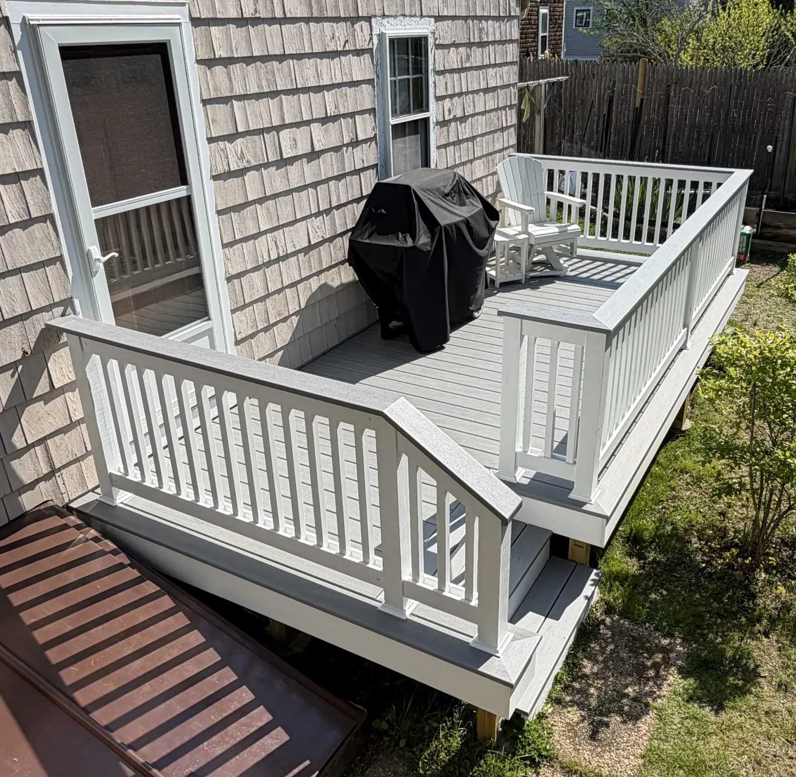 A view of a backyard deck featuring a white railing, a black grill covered with a protective cover, and a white outdoor chair. The deck is sunlit, adjacent to a house with shingle siding.