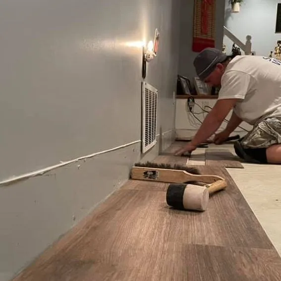 Contractor installing wood like vinyl flooring in gray room. Handle held brush broom and mallet laying on flooring.
