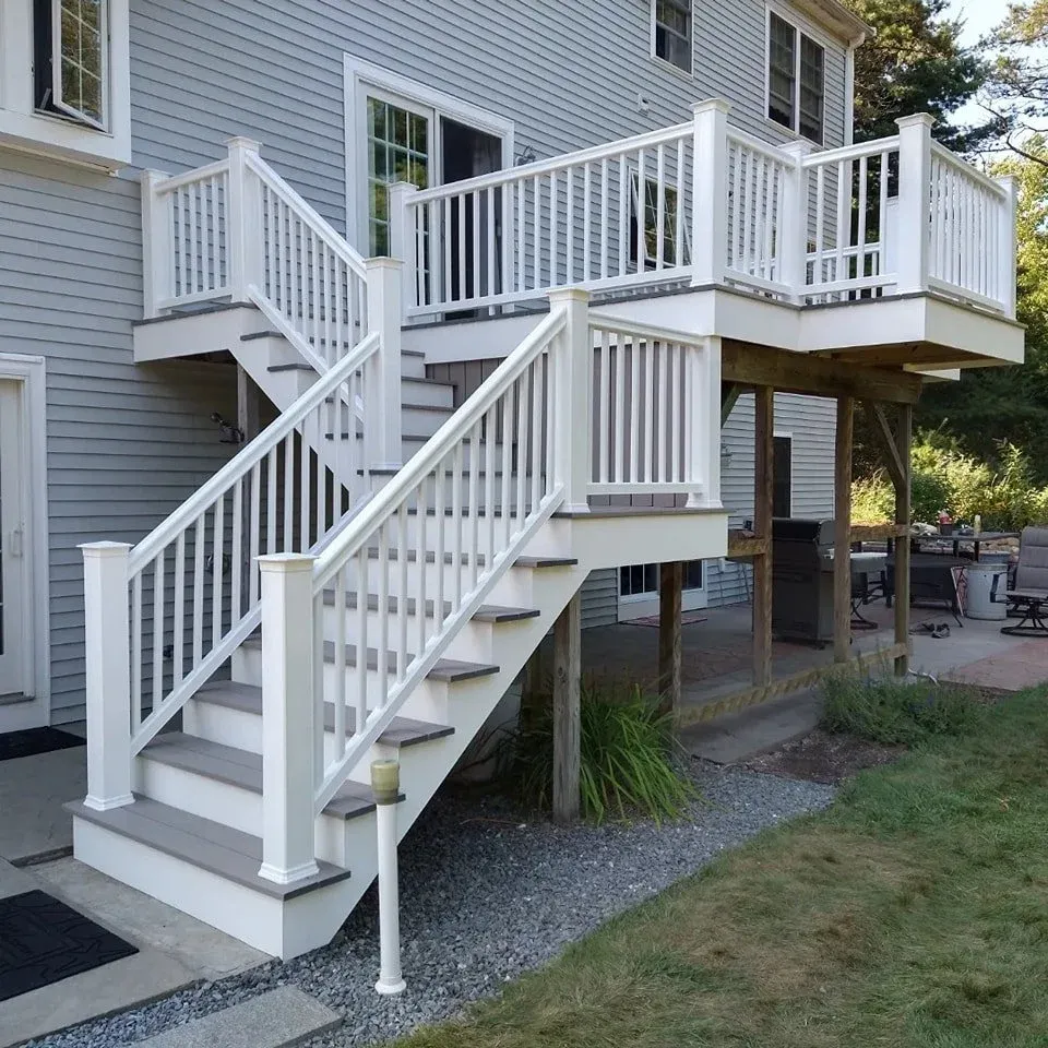 A white wooden staircase with railing leading up to a second-story deck attached to a gray house with horizontal siding.