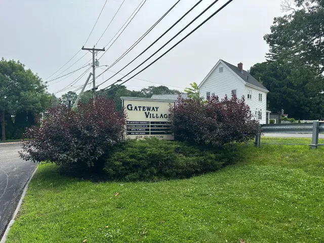 Neighborhood entrance sign by a road, bordered by shrubs and a white house under power lines.