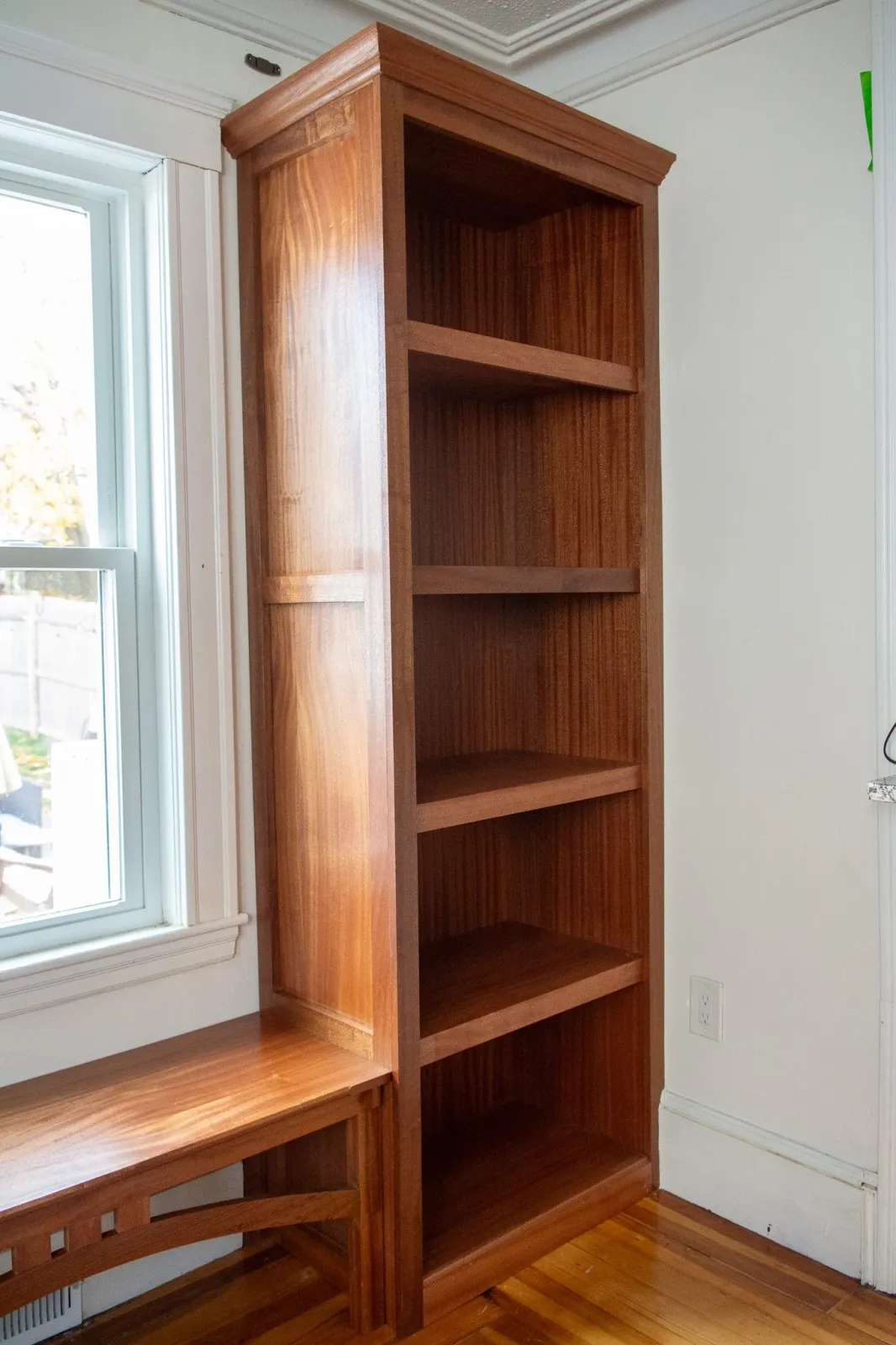 A tall wooden bookshelf with five shelves, positioned next to a window in a room with hardwood floors and white walls.