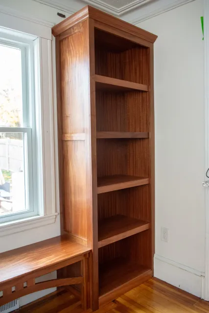 A tall wooden bookshelf with five shelves, positioned next to a window in a room with hardwood floors and white walls.