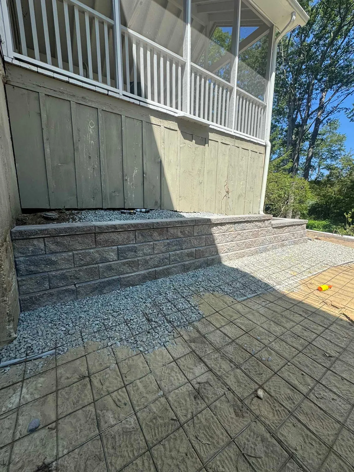 A new stone retaining wall sits beneath a porch, with gray gravel surrounding the base and wire mesh laid on the ground.