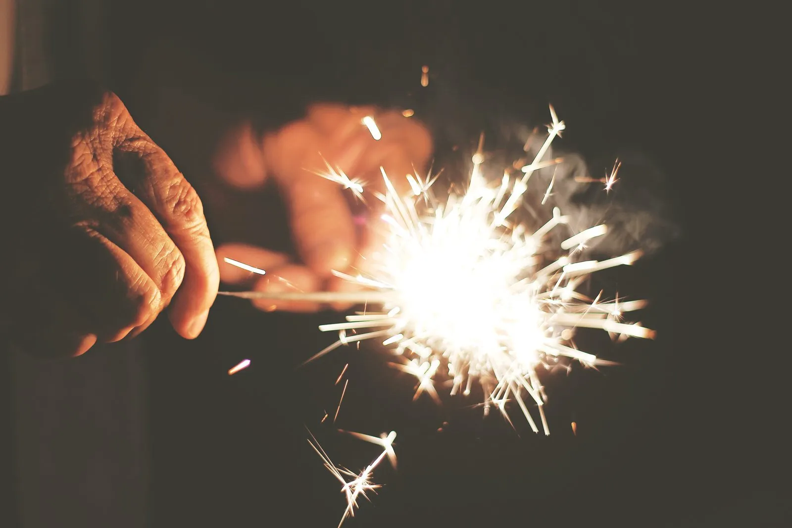 Person holding a lit sparkler, emitting bright sparks against a dark background.