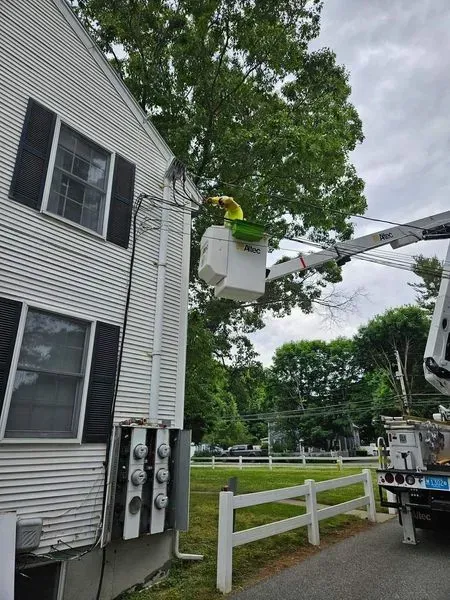 Electrician in a lift bucket trims tree branches near power lines attached to a white building.