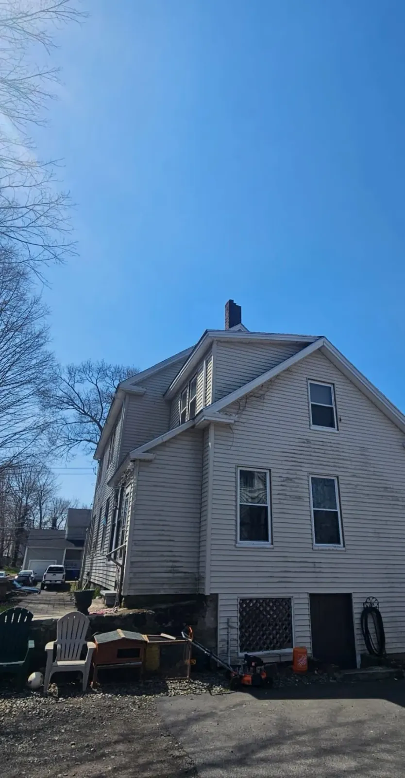 White two-story house with a steep roof and siding, viewed from the side under a clear blue sky.